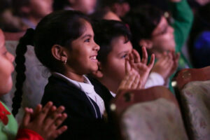 A young girl reacts to a performance with a smile and hands clapping.